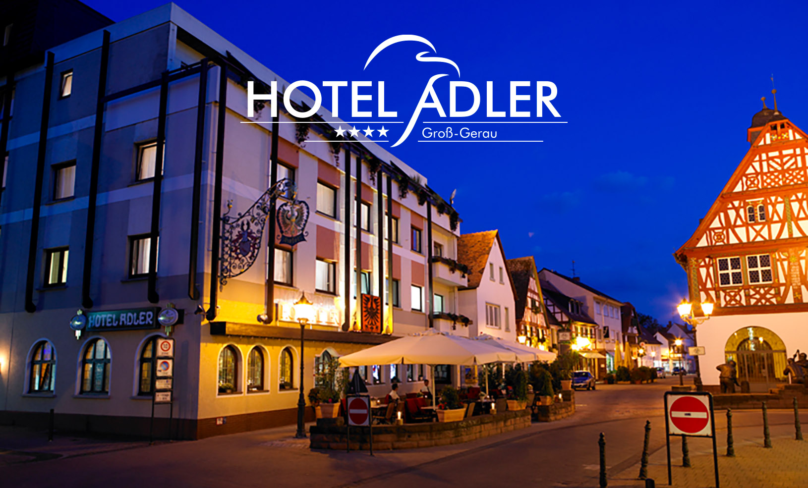Erleuchtete Straßenszene in der blauen Stunde mit Fachwerkhäusern und stimmungsvoller Straßenbeleuchtung im Sommer mit Schriftzug und Logo "Hotel Adler vier Sterne Groß-Gerau": Das Hotel Adler ist ein Eckhaus mit drei Obergeschossen. Davor befindet sich in einem verkehrsberuhigten Bereich ein Biergarten mit Sonnenschirmen. Auf der anderen Straßenseite steht das Historische Rathaus, ein altes Fachwerkhaus mit zwei Obergeschossen.
