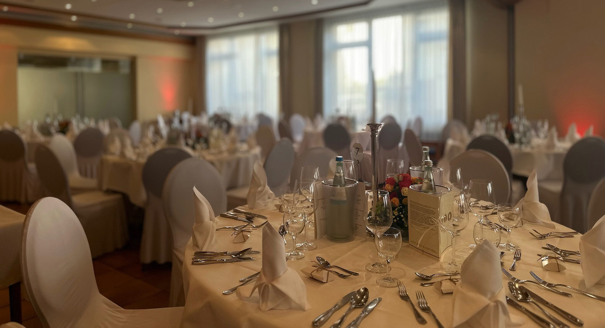 Festlicher Saal mit mehrere Runden Tischen für acht bis zehn Personen. Tischdecken, Servietten und Stuhlhussen sind aus Stoff. Es stehen Wasserflaschen in Flaschenkühlern, Menukarten und Blumenschmuck auf den Tischen. Das Besteckt wirkt hochwertig.