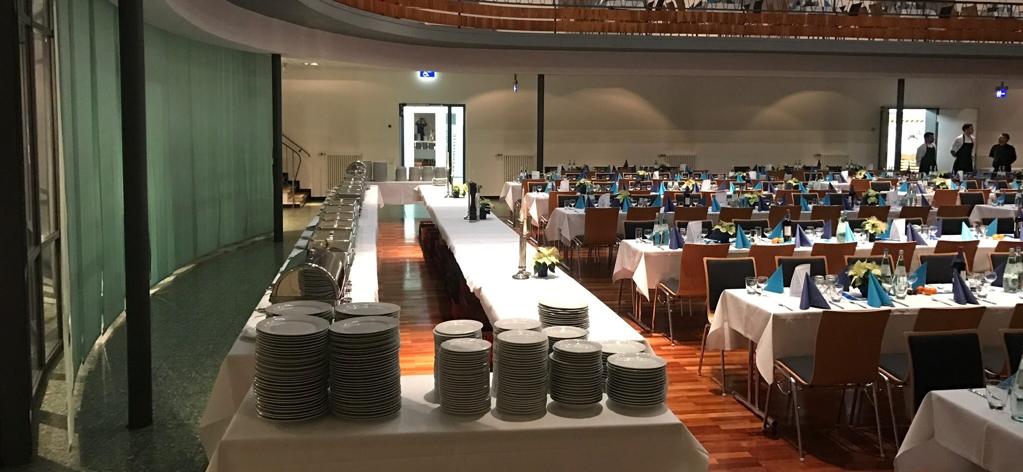 Stadthalle Groß-Gerau in Bankett-Bestuhlung mit Cateringbuffet: großer Saal mit Empore. In der Bildmitte ist ein Catering-Buffet aufgebaut. Das Parkett ist bestuhlt mit überwiegend langen Tischen, an denen man sich frontal gegenübersitzt. Auf und unter der Empore sind 6er Tische. Die Tische sind mit einer Stofftischdecke, Blumendekoration und farbigen Papierservietten gedeckt.