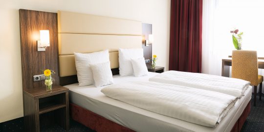 Hotel_Adler_Zimmer_218_0032_1E_2500px Comfort Doppelzimmer: Doppelbett mit jeweils einem Nachttisch an der Seite. Dort befinden sich jeweils auch Lichtschalter und Steckdosen. Vor dem Fenster steht ein Stuhl und ein Schreibtisch. Der Bodenbelag ist Teppich.
