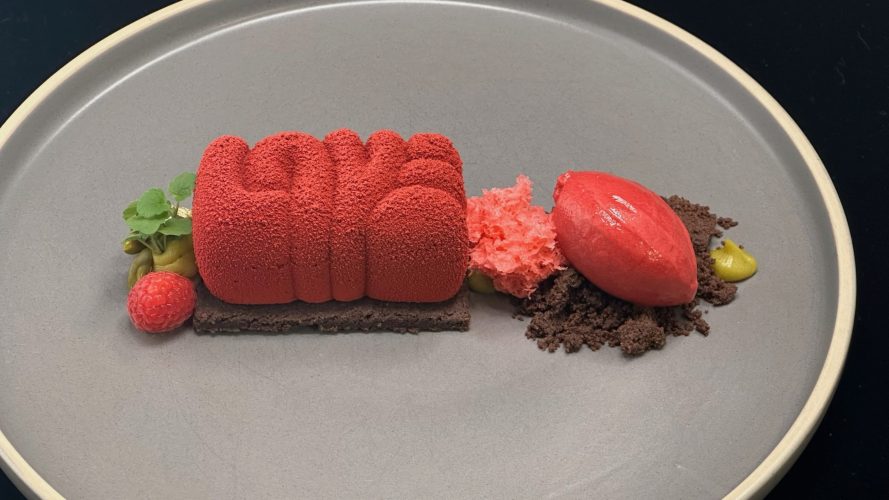 Menuvorschlag Nachtisch: Auf einem grauen Teller befindet sich ein roter Fruchtnachtisch auf einem Schokobett garniert mit Obst.