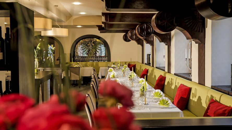 Restaurant im Hotel Adler: Es gibt Stühle und eine lange hellgrüne Bank zum Sitzen mit roten Kissen. Festlich gedeckte Tische mit grün-weißen Servietten, Kerzen und Blumenschmuck. Der Raum ist hell mit weißen Wändern und dunklem Holz. Im Vordergrund sieht man eine rote Blumendekoration.