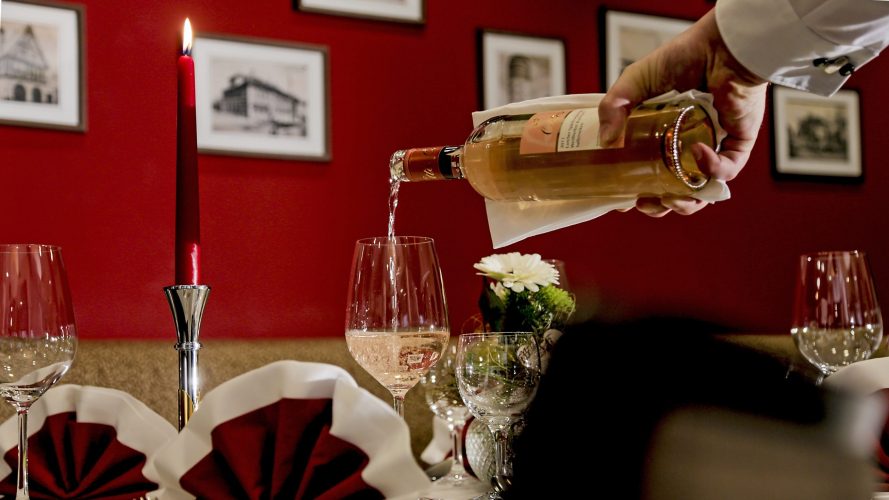 Festlich gedeckter Tisch vor einer roten Wand mit Bildern die historische Motive zeigen. Auf dem Tisch steht eine brennende rote Kerze, weiß-rote Servietten und Gläser. Eine Hand gießt Roséwein in ein Weinglas.