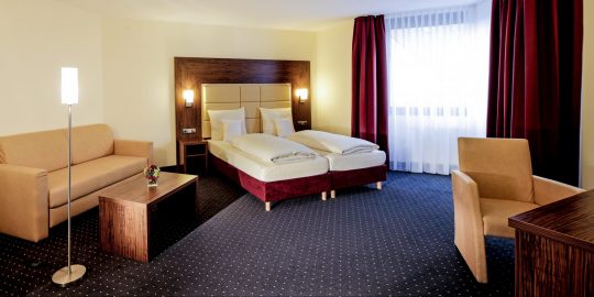 Zimmer 116 2 Junior Suite Doppelbett mit jeweils einem Nachttisch an der Seite. Dort befinden sich jeweils auch Lichtschalter und Steckdosen. Im Hintergrund ist ein großes helles Fenster. Im Vordergrund rechts steht ein Sessel und ein Schreibtisch. Im Vordergrundlinks stehen eine Couch, ein Couchtisch mit Blumenschmuck und eine Stehlampe. Der Bodenbelag ist Teppich.