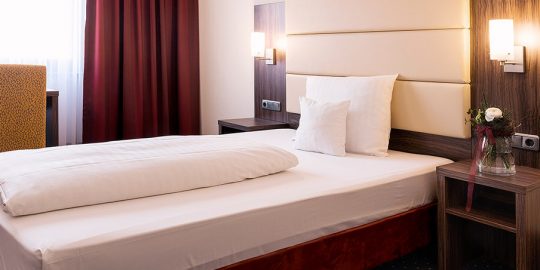 hotel-adler-zimmer-07 Comfort Einzelzimmer: Einzelbett mit jeweils einem Nachttisch an beiden Seiten. Dort befinden sich jeweils auch Lichtschalter und Steckdosen. Vor dem Fenster steht ein Stuhl und ein Schreibtisch. Der Bodenbelag ist Teppich.